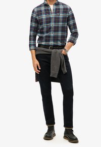 Multikleurige geruite overhemd met lange mouwen, donkere spijkerbroek, grijs vest om de taille geknoopt en zwarte veterschoenen.