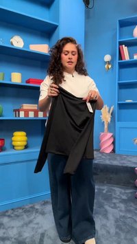 Frau mit lockigem Haar hält ein schwarzes Langarmshirt und steht in einem blauen Raum mit Regalen, die bunte Gegenstände und Bücher halten.