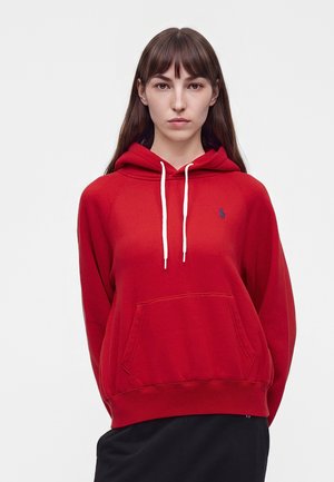Femme aux longs cheveux bruns portant un sweat à capuche rouge avec des cordons blancs et un petit logo bleu sur la poitrine, debout sur un fond blanc.
