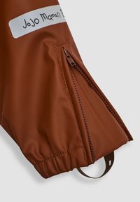 Tela impermeable marrón con un bolsillo con cremallera, puño elástico y una etiqueta que dice "JoJo Maman Bébé". Textura suave y mate, diseño práctico.