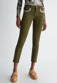 Pantalones color verde oliva con un ajuste ceñido, que presentan detalles de pedrería en los bolsillos delanteros y un corte ligeramente cropped por encima del tobillo. Tejido de textura suave.