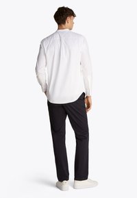 Camicia bianca a maniche lunghe con orlo curvo e polsini abbottonati, abbinata a pantaloni scuri e scarpe da ginnastica bianche, vista da dietro.