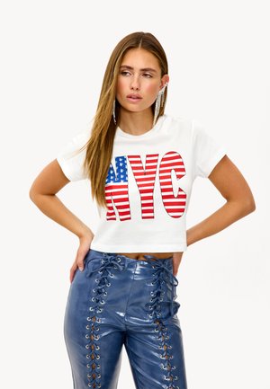 T-shirt blanc court avec du texte "NYC" en rouge et bleu, sur un motif de drapeau. Porté avec un pantalon en simili cuir bleu taille haute avec des côtés lacés.