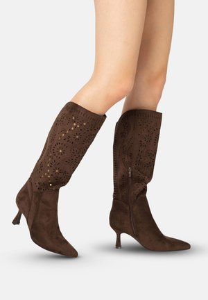 Brune ruskind knæhøje støvler med blomsterudskæringsmønstre, guldfarvede nitter, sidelukning med lynlås og spidse tæer på slanke hæle, båret på bare ben.
