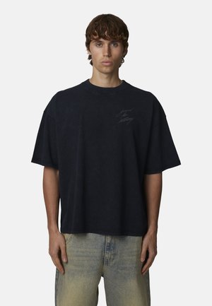 Giovane uomo con capelli castani che indossa una maglietta nera ampia con testo appena visibile e jeans chiari slavati, in piedi davanti a uno sfondo bianco uniforme.