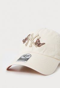 Krämfärgad baseballkepp med broderad NY-logga och fjärilsmotiv, justerbar rem och märkesetikett på vänstra framsidan.