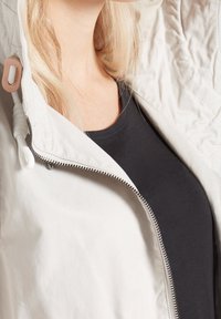 Giacca leggera beige con cappuccio imbottito, dotata di chiusura con zip e cordino sul colletto. Top nero sotto.