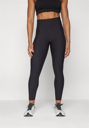 Leggings atléticos negros con cintura alta, con tejido texturizado y detalles de panel en diagonal. Combinados con zapatillas atléticas blancas.