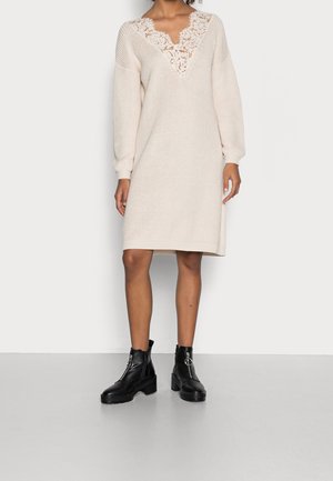 Mujer con un vestido de punto color crema con detalle de encaje en forma de V en el escote y mangas largas, combinado con botas negras gruesas de tobillo con anillas en la cremallera.