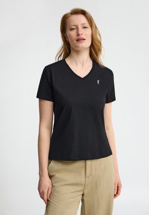 Femme à la peau claire et aux cheveux roux portant un T-shirt noir à col en V avec un petit logo blanc et un pantalon beige, debout devant un fond uni.