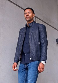 Mørkeblå læderjakke med ståkrave, lynlåse-lommer, lagdelt design, parret med en mørk skjorte og blå jeans.