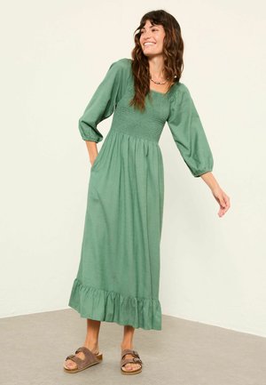 Grünes, langärmeliges Maxikleid mit gerafftem Oberteil, gerafftem Saum und weicher Textur. Kombiniert mit braunen Slide-Sandalen.