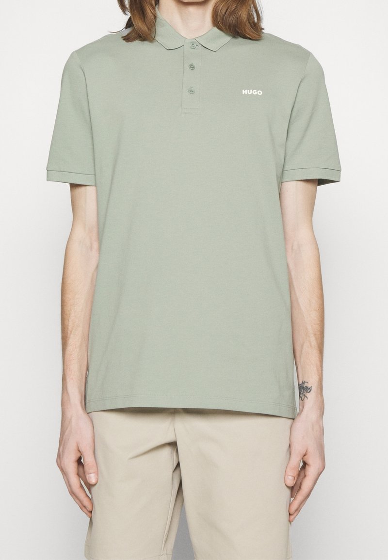 Polo manches courtes vert clair avec trois boutons et logo blanc "HUGO" sur la poitrine, porté avec un short beige.