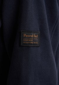 Granatowy sweatshirt z prostokątną czarną metką z brązowym napisem "Petrol Ind Denim Driven" oraz detalami przeszycia.