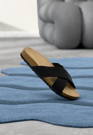 Enkelt sort slidesandal med korkfodaftryk, der hviler på et tekstureret blåt tæppe, sløret stor peber i baggrunden.