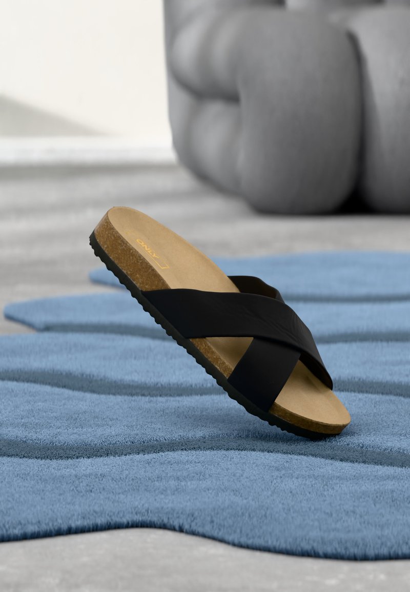 Une sandale noire à semelle en liège posée sur un tapis bleu texturé, un gros poivron flou en arrière-plan.