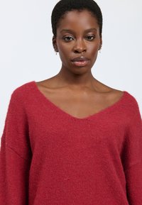 Maglione rosso a V in tessuto morbido e strutturato; vestibilità ampia con spalle scese e dettagli a coste sul collo e sull'orlo.
