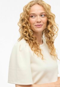 Haut à manches courtes de couleur crème avec un col rond et des manches bouffantes, confectionné dans un tissu lisse et léger.