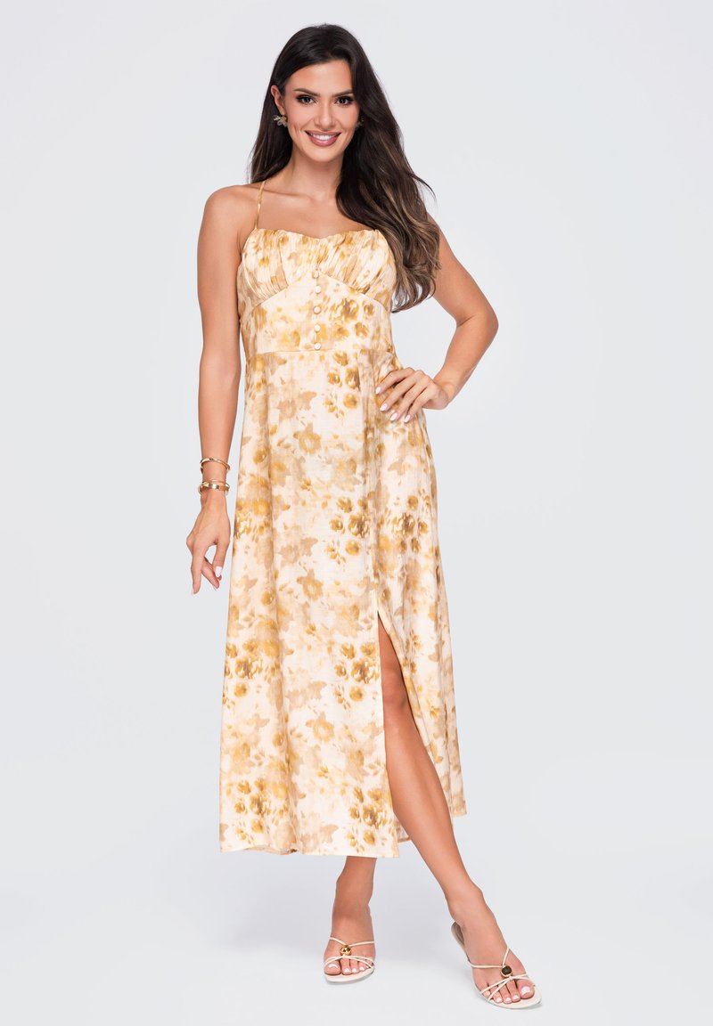 Femme en robe midi jaune fleurie sans manches avec fente latérale, souriante, debout avec une main sur la hanche et portant des talons blancs à brides.