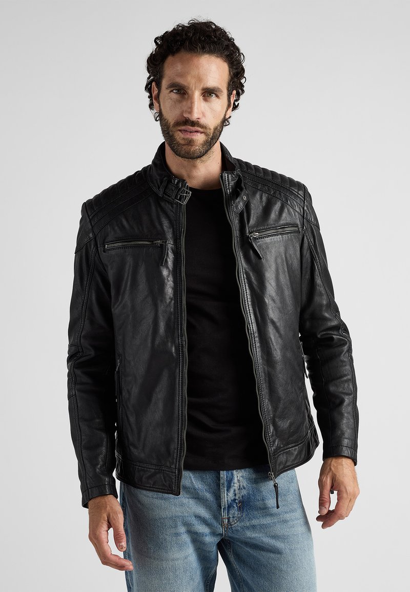 Schwarze Leder-Motorradjacke mit Reißverschlusstaschen, gerippten Schulterdetails und Stehkragen. Getragen über einem schwarzen Shirt und blauen Jeans.