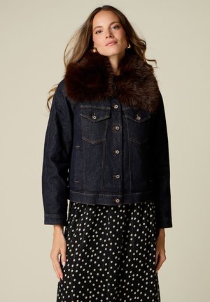 Donna con giacca di denim scuro con grande collo di pelliccia marrone scuro e gonna nera con pois bianchi, in piedi davanti a uno sfondo semplice.
