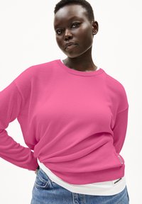Helles pinkes Sweatshirt mit rundem Halsausschnitt und gerippten Bündchen, über ein weißes Hemd gezogen. Besitzt ein sichtbares Markenetikett in der Nähe des Saums.