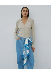 Beżowy kardigan prążkowany z dekoltem w serek i guzikami, połączony z niebieskimi dżinsami o wysokim stanie i wzorzystą niebiesko-białą chustą.