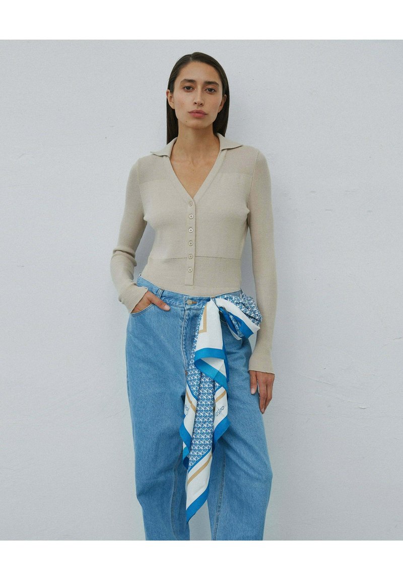 Beżowy kardigan prążkowany z dekoltem w serek i guzikami, połączony z niebieskimi dżinsami o wysokim stanie i wzorzystą niebiesko-białą chustą.