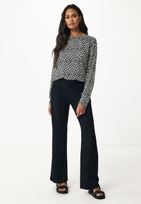 Top a maniche lunghe a motivi bianco e nero abbinato a pantaloni neri a vita alta svasati e scarpe scivolate nere, su uno sfondo semplice.
