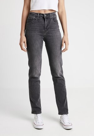 Frau trägt hoch taillierte, dunkelgraue Slim-Fit-Jeans, weißes, kurz geschnittenes geripptes Tanktop und weiße Sneaker, steht vor einem schlichten Hintergrund.