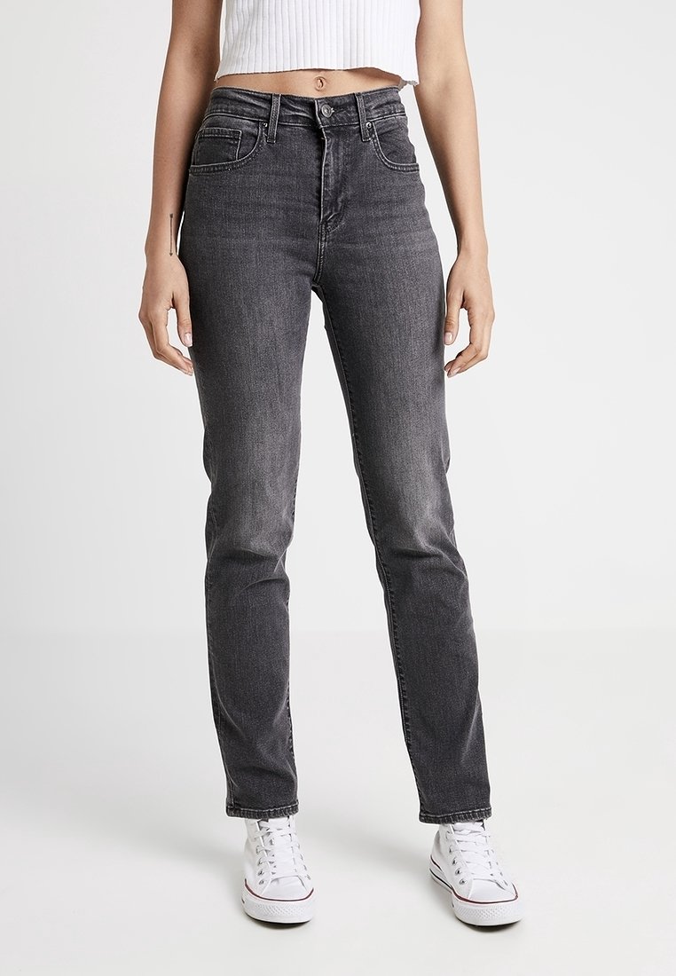Femme portant un jean slim taille haute gris foncé, un débardeur blanc court en côtes et des baskets blanches, debout devant un fond uni.