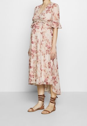 Femme enceinte portant une robe midi rose pâle à fleurs avec des manches volantées et des sandales plates marron à lacets, debout devant un fond uni.