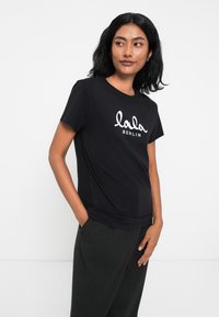 Melnā T-kreklā, kas izgatavota no mīksta auduma, ir balti slīpi ierakstīta teksts "lala BERLIN". Īsas piedurknes un brīvs piegriezums.