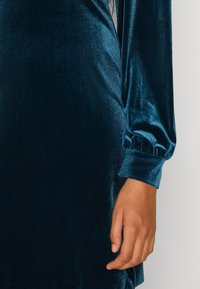 Tyrkysové sametové šaty s dlouhými, nabíranými rukávy s elastickými manžetami a hladkou, lesklou texturou. Detailní pohled na rukáv a ruku.