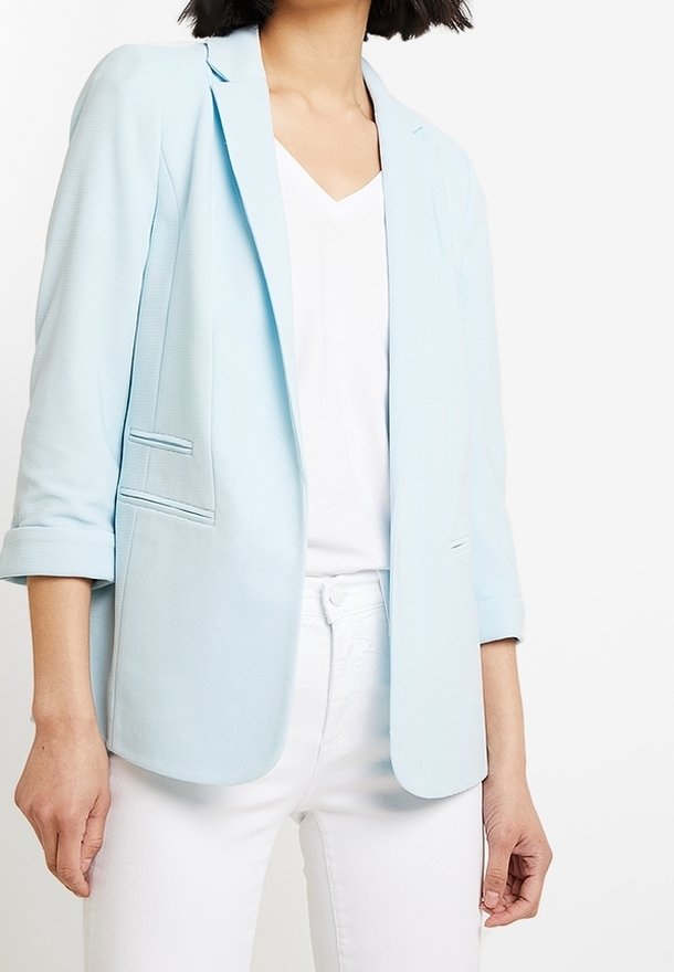 Femme portant un blazer bleu clair par-dessus un haut blanc à col en V et un pantalon blanc, debout devant un fond blanc uni.