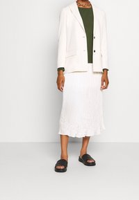 Vit kavaj över en grön topp, kombinerad med en vit plisserad midi-kjol och svarta glidsandaler. Slät tyg med synliga texturdetaljer.