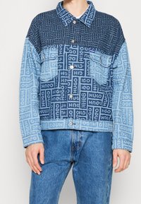 Kurtka dżinsowa z wzorem patchwork w ciemnym i jasnym odcieniu niebieskiego, z nadrukiem „HUGO” na całej powierzchni. Dwie kieszenie na piersi i zapięcie na guziki.