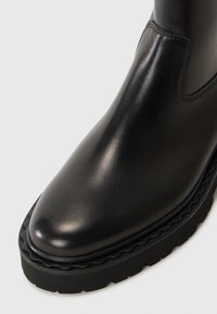 Bottine en cuir noir avec une finition lisse, bout rond et semelle en caoutchouc crantée. Présente des coutures minimales et un panneau élastique sur le côté.