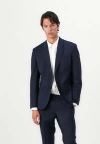 Costum bleumarin realizat pe comandă, cu două butoane, rever cu vârf, și două buzunare frontale, asortat cu o cămașă albă cu nasturi, textură netedă.