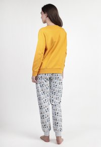Una mujer joven está descalza, de pie y mirando hacia la izquierda, vistiendo una camiseta amarilla de manga larga y pantalones de pijama grises con estampado de personajes de dibujos animados.