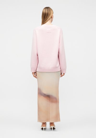 Γυναίκα με μακριά ξανθά μαλλιά φοράει ένα ανοιχτό ροζ πουλόβερ, μια μακριά φούστα με μπεζ και καφέ σχέδιο και λευκά παπούτσια με τακούνι, στέκεται γυρισμένη με την πλάτη.