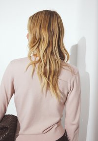 Maglione rosa chiaro con maniche lunghe e vita aderente, abbinato a una borsa a tracolla marrone intrecciata. Capelli testurizzati che scendono lungo la schiena.