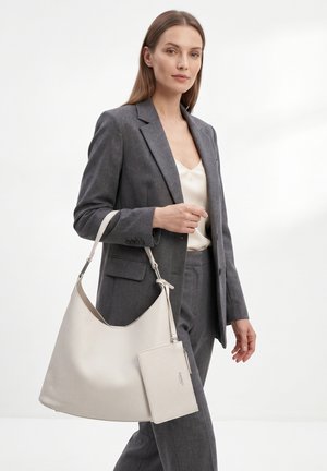 Vrouw in grijs broekpak die een grote off-white leren handtas met bijpassend tasje draagt, staand tegen een effen witte achtergrond.