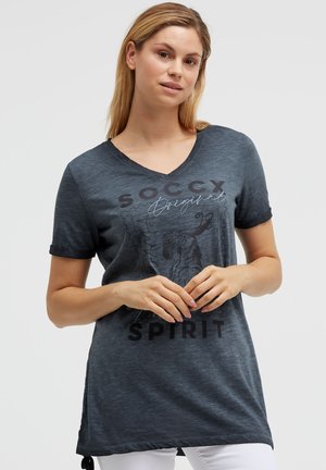 Woman with blonde hair wearing a dark gray V-neck T-shirt with text "SOCCX Original Spirit" and a graphic, standing against a plain background.