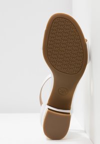 Sandale avec des brides blanches et une semelle marron montrant un motif répétitif du logo "MK" en relief, talon surélevé, placée sur fond blanc.