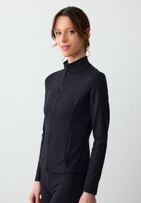 Giacca sportiva nera aderente con colletto alto e zip completa, realizzata in materiale morbido ed elastico. Presenta tasche laterali e maniche lunghe.