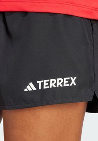 Juodi sportiniai šortai, pagaminti iš tekstūruotos medžiagos, su raudona juosta, ant kurios aiškiai matomas baltas „TERREX“ logotipas apatinėje pusėje.