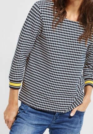 Femme portant un haut à manches longues à pois noir et blanc avec des poignets rayés jaunes, associé à un jean bleu, debout avec une main dans la poche.