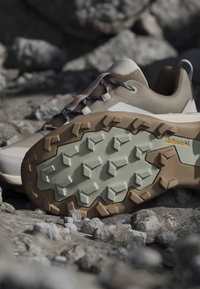 Chaussure de randonnée avec semelle robuste marron et beige à crampons profonds, posée sur un terrain extérieur rocheux.
