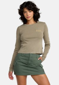 Langemouwige, olijfgroene cropped top met gouden logo, gecombineerd met een groene minirok in cargostijl met zakken en stiksels.